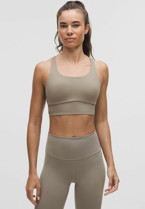 Tan sports-bh med brede stropper og en glat tekstur, kombineret med matchende højttaljede leggings, begge lavet af strækbart, tætsiddende materiale.