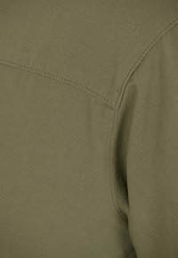 Vihreä oliivinvärinen kangas, pehmeä pinta, näkyvät saumat; lähikuva vaatteen olkapäästä, joka näyttää sauman yksityiskohtia ja hienoita kudoksia.