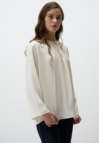 Blusa color crema con scollatura rotonda, maniche lunghe e una sottile texture, dotata di un dettaglio a chiave sulla parte anteriore.