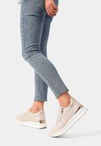 Sneaker beige chiaro con tomaia in pelle liscia, zip decorativa e dettagli metallici, abbinati a jeans skinny blu.