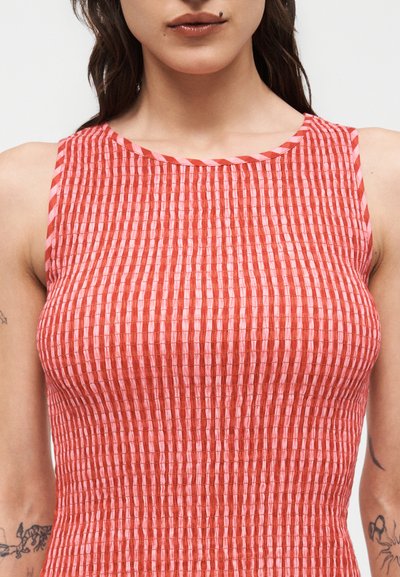 Κοντινό πλάνο μιας γυναίκας που φοράει αμάνικο φόρεμα με υφή, με ρίγες σε κόκκινο και ροζ χρώμα, με ορατά τατουάζ και στα δύο της χέρια.