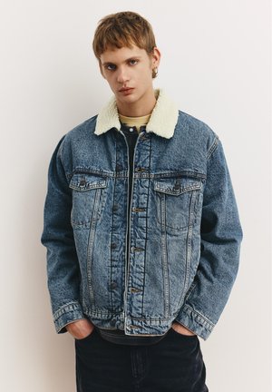 Jeune personne aux cheveux courts portant une veste en denim bleue avec un col sherpa blanc, les mains dans les poches, debout devant un fond blanc uni.