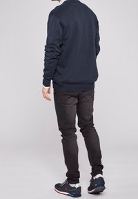 Man som bär en mörkblå tröja, svarta jeans och marinblå sneakers med vita sulor, står med ryggen vänd mot kameran mot en ljus bakgrund.