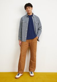 Fiatal férfi áll fehér fal előtt, csíkos szürke farmeringben, sötétkék pólóban, barna nadrágban és fehér cipőben kék fűzőkkel.