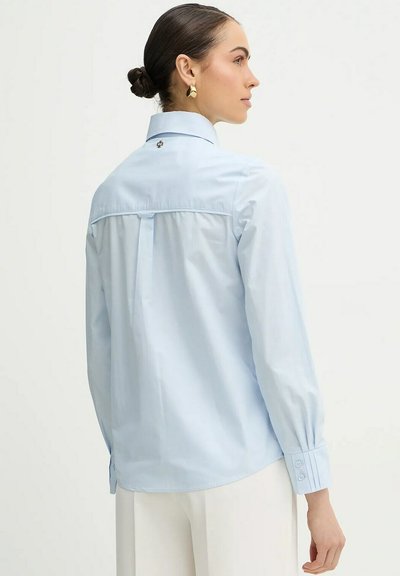 Donna con i capelli raccolti in uno chignon, che indossa orecchini d'oro, una camicia celeste a maniche lunghe con bottoni e pantaloni bianchi, vista da dietro e di profilo.