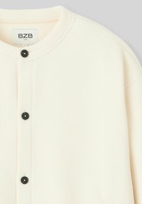 Veste beige à boutons en tissu texturé, avec un col rond, des boutons noirs et des accents côtelés. Design simple et fonctionnel.