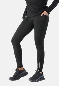 Leggings noirs en tissu lisse et extensible avec poches latérales et accents réfléchissants. Associés à des chaussures de sport sombres pour un look sportif.