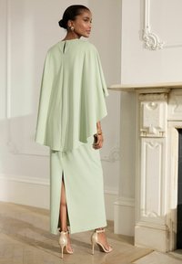 Robe cape vert pâle avec une fente haute à l'arrière, associée à des sandales à talons dorés. Tissu lisse avec des détails de design minimalistes.