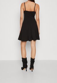 Kvinna i en svart ärmlös utsvängd klänning och svarta slappa högklackade stövlar som visas från ryggen mot en neutral bakgrund.