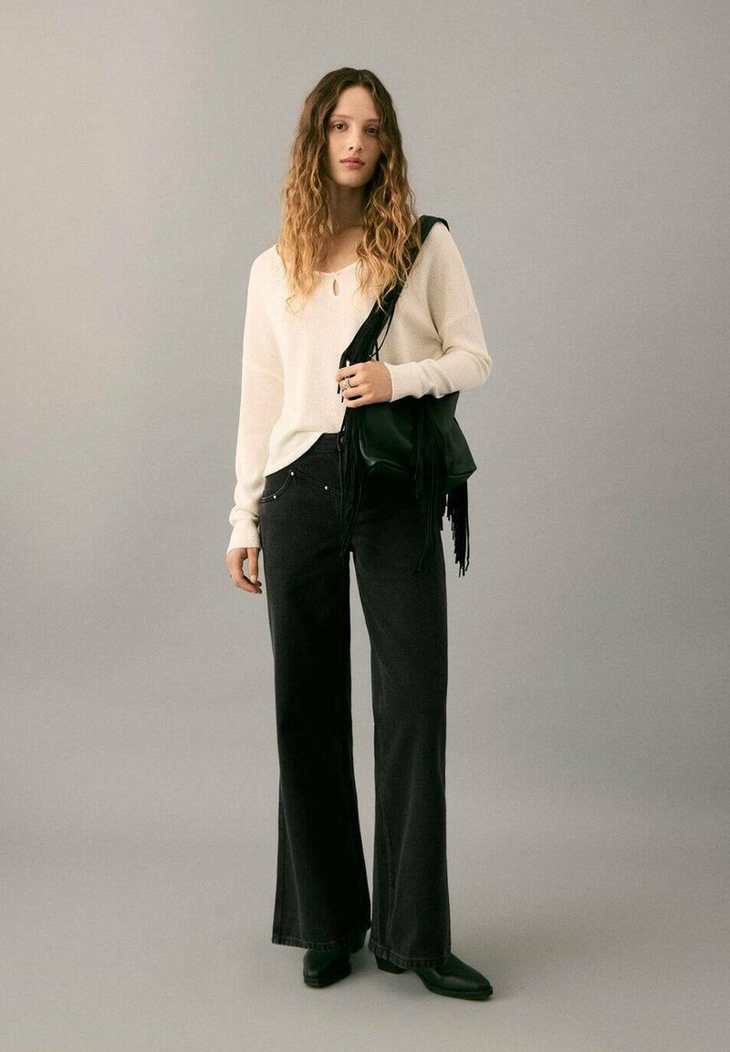 Jeune femme aux cheveux ondulés portant un pull crème, un pantalon noir à jambes larges, des bottes noires, et tenant un sac d'épaule à franges noir.