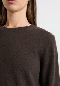 Pull en laine marron avec un col bateau côtelé, mettant en avant une texture douce et une silhouette cintrée, superposé sur un haut rose.
