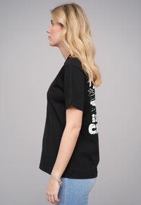 Black cotton t-shirt with short sleeves, featuring white graphic text on the back. Model wears light blue jeans and a bracelet.