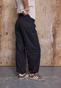 Pantalon noir, léger et ample avec une taille smockée, associé à des baskets marron ornées de rayures blanches. Tissu texturé et coupe décontractée.