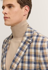 Blazer mit Karomuster in Beige, Marineblau und Creme, kombiniert mit einem hellbeigen Rollkragenpullover. Texturiertes Material und detaillierte Knöpfe.