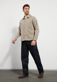 Veste texturée beige avec un col et une fermeture à boutons, dotée de deux poches avant, assortie à un jean bleu foncé et des chaussures marron.