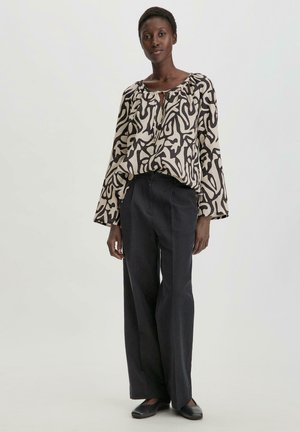 Blusa a maniche lunghe beige con un motivo astratto nero, caratterizzata da un collo rotondo e una vestibilità comoda. Abbinata a pantaloni neri a vita alta.