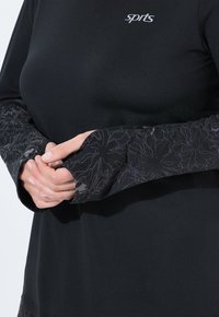 Zwarte sporttop met een aansluitend lichaam, voorzien van lange mouwen in een textuurpatroon van bloemen in donkere tinten en een logo op de borst.