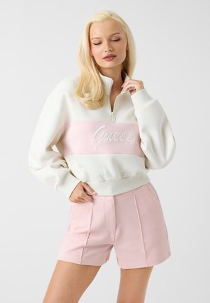 Donna con lunghi capelli biondi indossa una felpa Guess bianca e rosa con pantaloncini rosa, in piedi contro uno sfondo chiaro e uniforme.