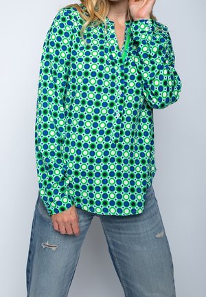Person iført en green, blå og hvid mønstret skjorte med knapper og blå revnede jeans, der poserer med den ene hånd hævet nær ansigtet.