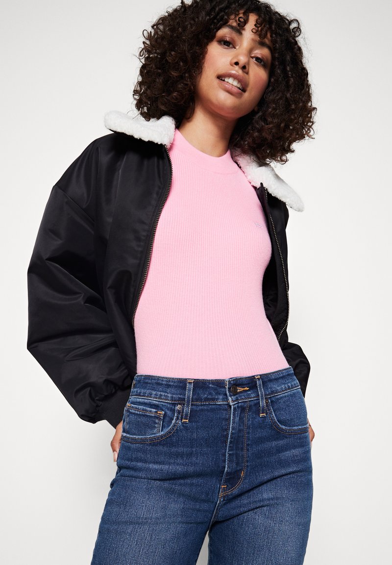 Giacca bomber nera con colletto in pelliccia sintetica bianca, top rigato rosa e jeans in denim blu. Caratterizzata da chiusura con cerniera e design classico a cinque tasche.