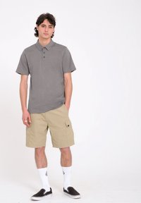 Polo gris avec trois boutons, shorts cargo beige avec poches latérales, chaussettes blanches et chaussures noires à enfiler. Design simple et décontracté.