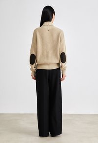 Vrouw met lang, donker haar die een beige gebreide trui met zwarte elleboogpatches en ruime zwarte broek draagt, staande met de rug naar de kijker gekeerd.