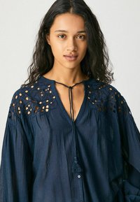 Blusa blu navy con dettagli ricamati a taglio sulle spalle, scollo a V e chiusura con cordoncino. Tessuto leggero con una vestibilità rilassata.