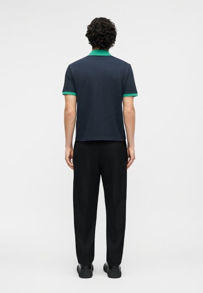 Άνδρας με σγουρά μαλλιά στέκεται με την πλάτη, φορώντας μπλε μπλούζα πόλο με πράσινο γιακά και μανσέτες, μαύρο παντελόνι και μαύρα παπούτσια.