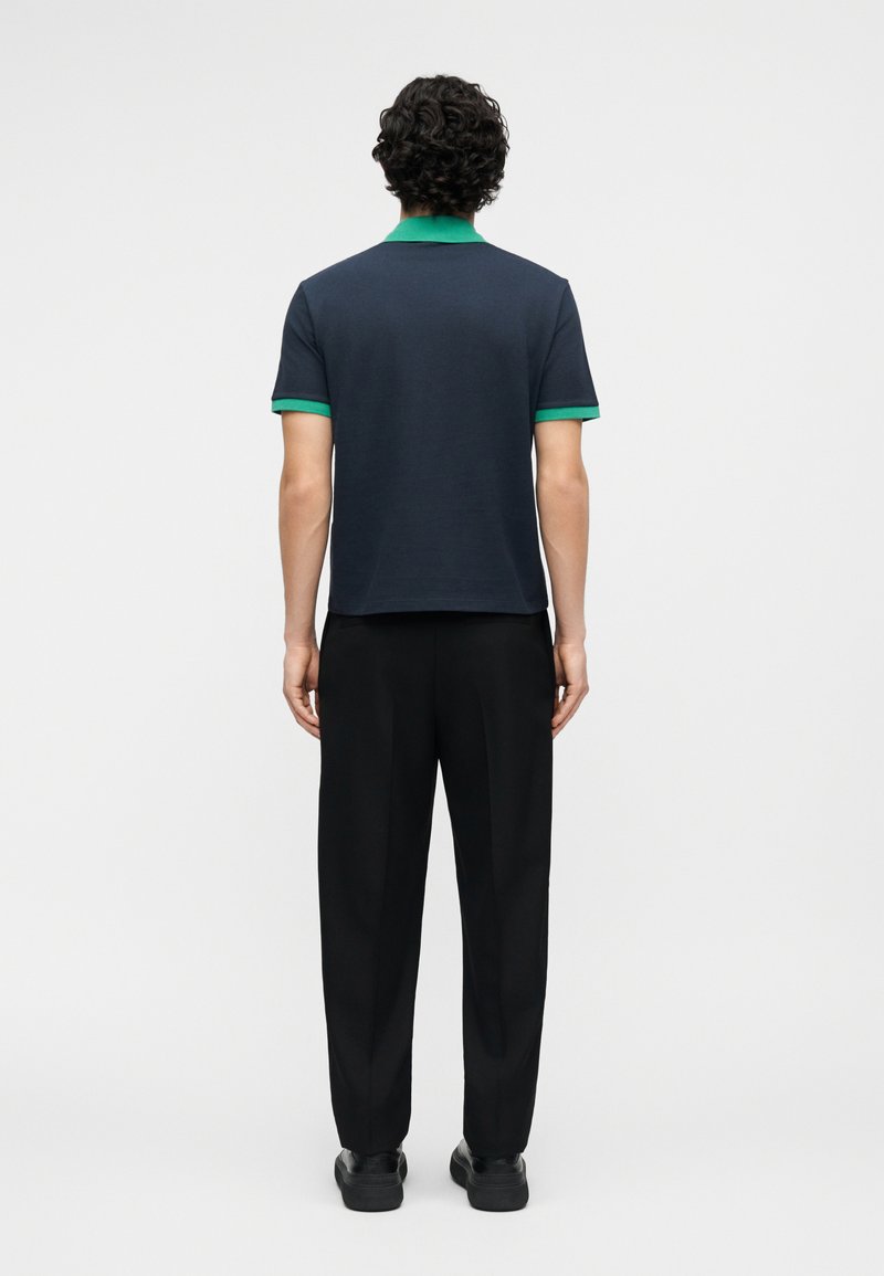 Man met krullend haar staande met zijn rug naar de camera, gekleed in een marineblauw poloshirt met groene kraag en manchetten, zwarte broek en zwarte schoenen.