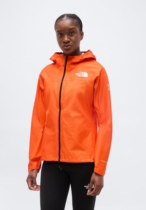 Γυναίκα που φοράει ένα φωτεινό πορτοκαλί μπουφάν με κουκούλα The North Face με μαύρο φερμουάρ και μαύρα κολάν, πάνω σε απλό φωτεινό φόντο.