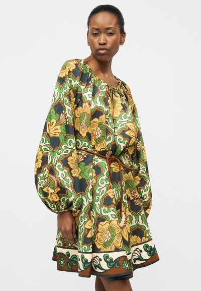 Γυναίκα που φοράει ένα φαρδύ, μακρυμάνικο φόρεμα με πράσινο, κίτρινο και καφέ λουλουδάτο και γεωμετρικό μοτίβο, που δένεται στη μέση με μια λεπτή ζώνη.