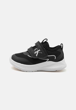 Black sneaker with mesh material, white accents, and Velcro strap. Features a rounded toe and textured sole for traction. Logo displayed on strap.