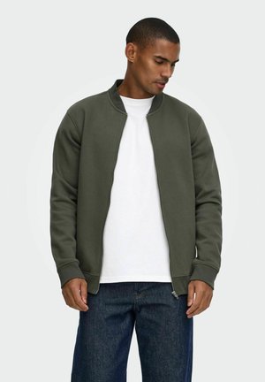 Mann trägt eine offene dunkelgrüne Jacke mit Reißverschluss über einem weißen T-Shirt und blauen Jeans, blickt nach unten vor einem einfarbigen hellen Hintergrund.