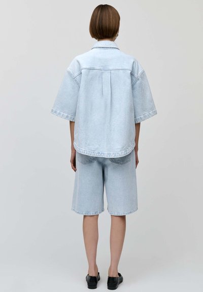 Camisa de denim azul claro de gran tamaño con mangas cortas y pliegue en la parte posterior, combinada con pantalones cortos hasta la rodilla. Ambos tienen un acabado desgastado y texturizado.