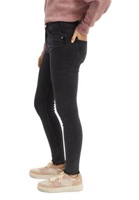 Vaqueros negros ajustados con una textura suave, que cuentan con bolsillos frontales y herrajes metálicos. Combinados con zapatillas de color claro.