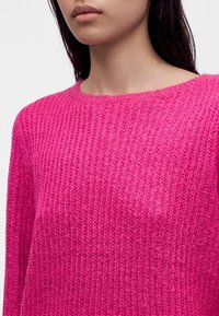Jeune personne portant un pull tricoté rose vif à col rond, visible du cou jusqu'au milieu du torse.