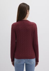 Burgunderfarbener Kabelstrickpullover mit gerippten Bündchen und Saum, der ein strukturiertes Muster aufweist. Getragen mit hellblauen Jeans, von hinten betrachtet.