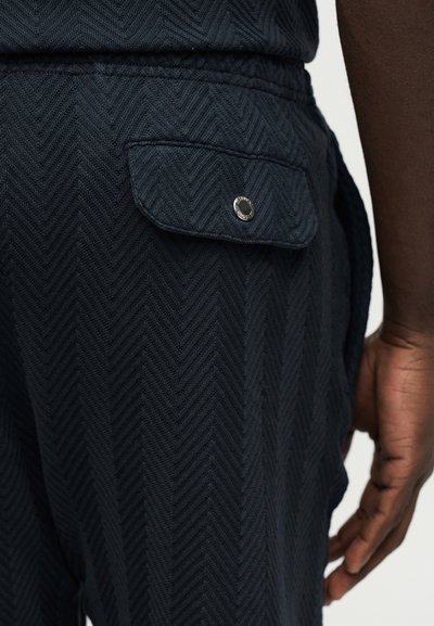 Gros plan sur un pantalon à motif chevrons sombre avec une poche à rabat fermée par un bouton en métal, porté par une personne à la peau foncée.
