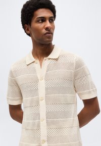 Άνδρας με κοντά σγουρά μαλλιά που φοράει μπεζ πλεκτό πόλο με κοντά μανίκια και ριγωτό ανοιχτό πλέγμα.