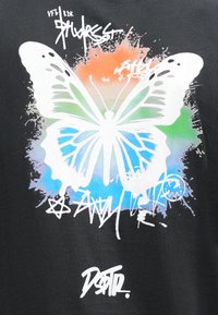 Schwarzes T-Shirt mit großem weißen Schmetterlingsgrafik, ergänzt durch blaue, grüne und orangefarbene Farbspritzer sowie bunten Graffiti-Text und Symbole.