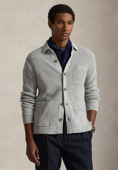 Man draagt een lichtgrijs gebreid vest met knopen over een marineblauw overhemd met een polkadot sjaal en donkere, fijn gestreepte broek, staand tegen een effen achtergrond.