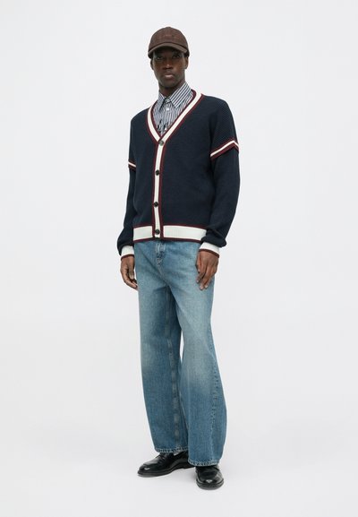 Cardigã de malha azul-marinho com riscas brancas e bordô, fechamento com botões, usado sobre uma camisa riscada, combinado com jeans azul claro e sapatos pretos.