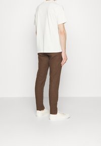 Bílý bavlněný tričko s krátkým rukávem a slim-fit hnědé lněné kalhoty, doplněné krémovými teniskami, zachyceno ze zadní strany na bílém pozadí.