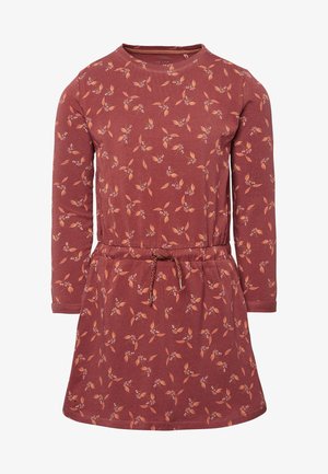 Barn i en rød blomstrede langærmet kjole, orange leggings, lyserøde sokker og lyserøde sneakers, der står sideværts med hånden i kjolens lomme.