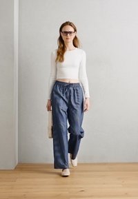 Model oblečena v prilagojen vrhnji beli dolg rokav in široke modre hlače z vzorcem ter elastičnim pasom, nosi svetlo torbo.