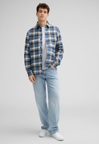 Karierte Button-Up-Hemd in Blau und Creme, kombiniert mit hellblauen Jeans und weißen Schuhen. Lange Ärmel, lockere Passform und dezente Textur.