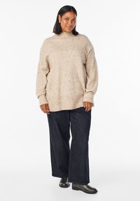 Beige Strickpullover mit gesprenkelt Muster, hohem Kragen und lockerer Passform, kombiniert mit dunkelblauen Denim-weitleg-Hosen und braunen Stiefeletten.
