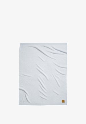Lyseblåt strikket tæppe med ribkant og et lille brunt mærke i nederste højre hjørne, liggende fladt med synlige folder i stoffet.