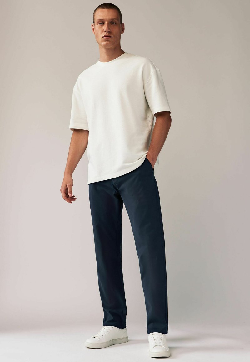 Witte T-shirt met korte mouwen en een relaxte pasvorm, gecombineerd met donkerblauwe broek en witte sneakers. Het T-shirt heeft een ronde hals.