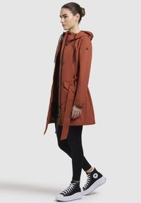 Roest-oranje waterdichte jack met capuchon, een aangesnoerde taille en zakdetails. Draagt over een zwarte top en gecombineerd met zwarte leggings en sneakers.